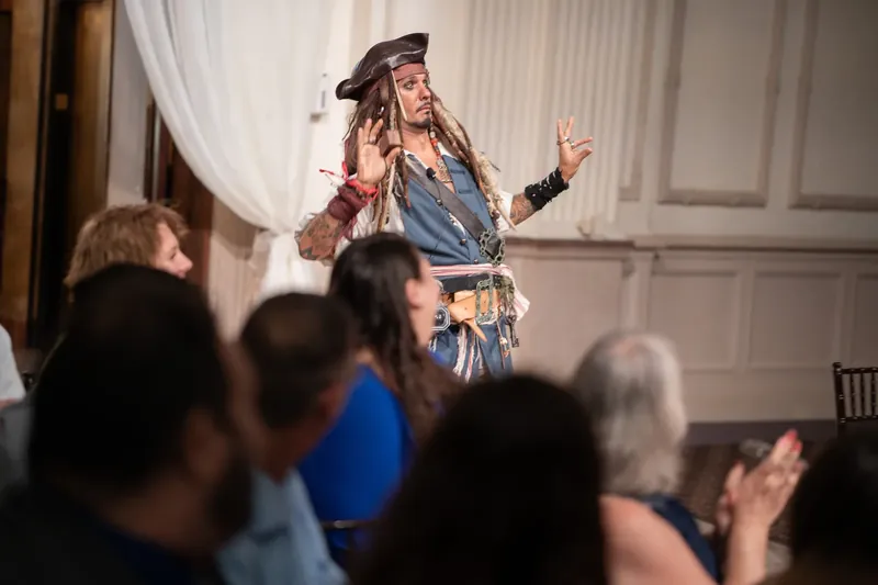 A performer in a pirate costume entertains wedding guests, gesturing expressively with arms spread wide.