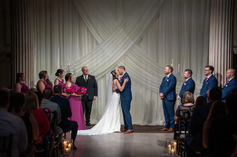 John and Jessica stand together at the altar beneath draped white fabric, flanked by wedding party members and lit candles.