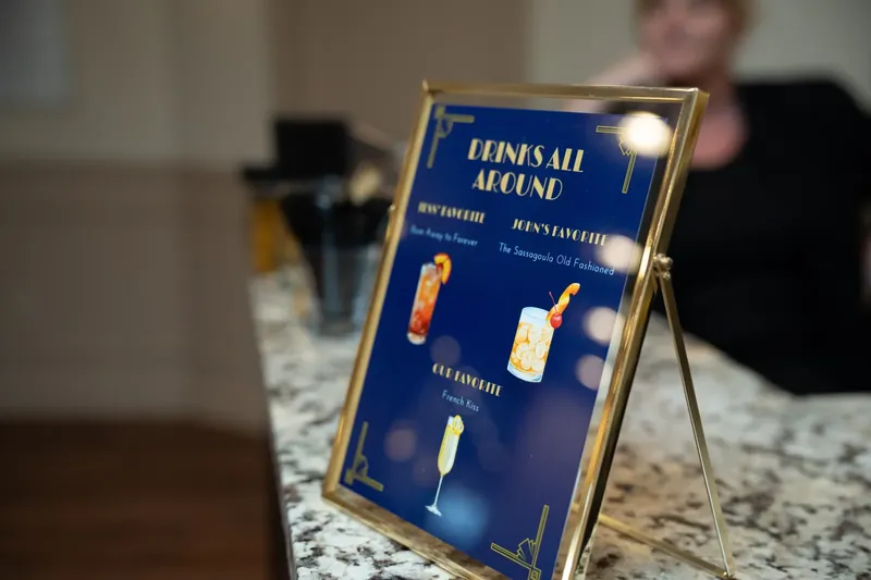 A gold-framed sign on a marble table lists cocktail options in blue and gold lettering at the reception.