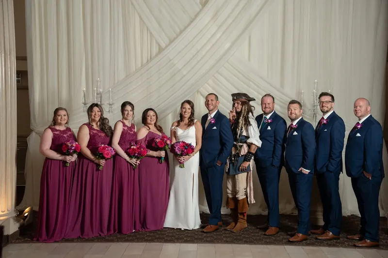 Jessica and John pose with their wedding party of five bridesmaids in burgundy and five groomsmen in navy against draped fabric.