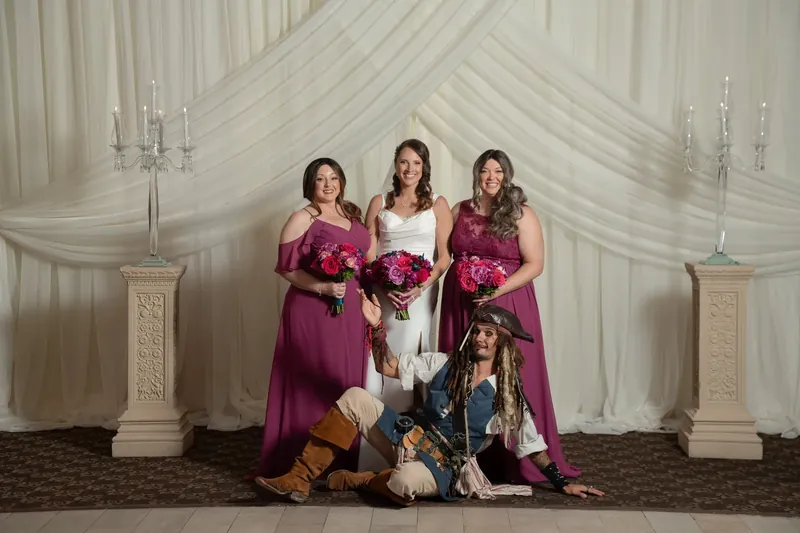 Jessica in a white gown stands with two burgundy-dressed bridesmaids holding red bouquets, a musician seated before them.