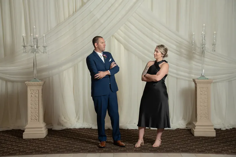 A man in a navy suit and a woman in a black dress stand facing each other against the draped white backdrop.