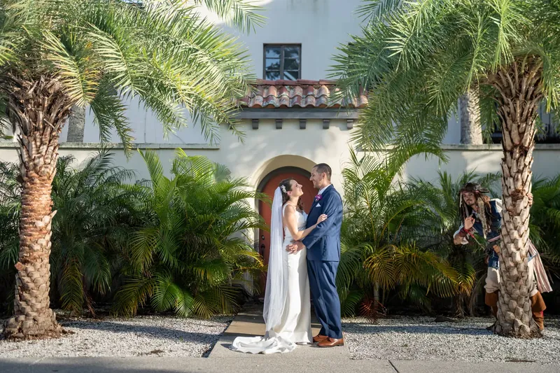 Jessica and John embrace and kiss under palm trees in front of a white stucco building with a wooden archway.