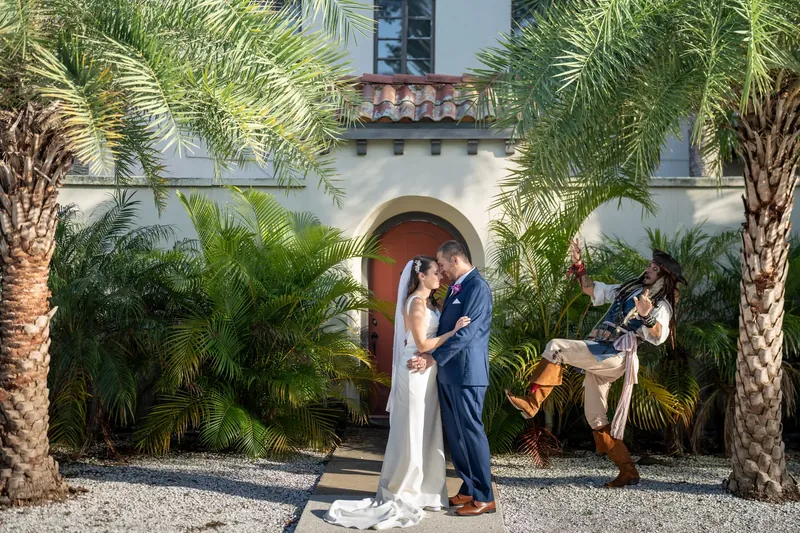The couple kisses while a costumed performer plays guitar nearby, framed by tall palms and the historic white facade.