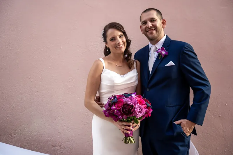 Jessica clutches a vibrant pink bouquet while standing beside John in a navy suit against a pale pink wall.