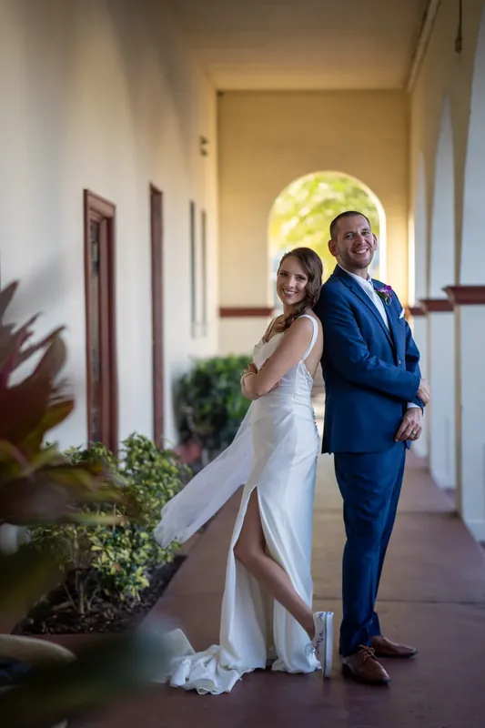Jessica and John stand back-to-back in an arched corridor, Jessica in a white gown and John in a navy suit.