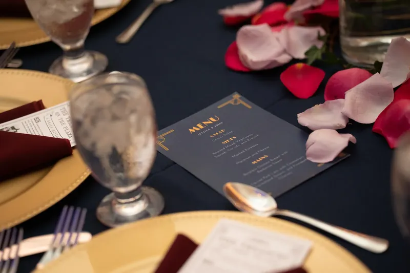 A dark blue menu card sits on a black tablecloth surrounded by water glasses, rose petals, and place settings.