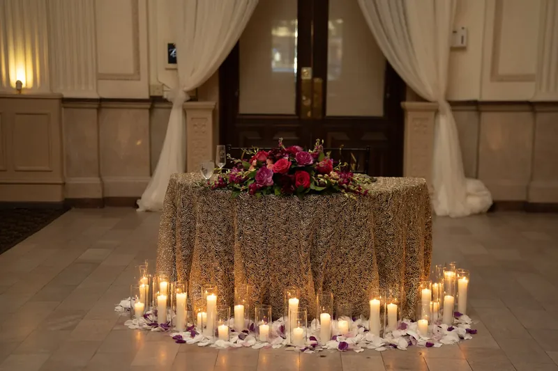 A gold-sequined sweetheart table with red florals and sheer draping is surrounded by lit candles.