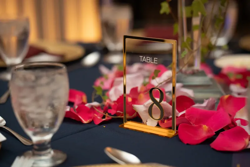 A gold acrylic table number "8" sits amid scattered red petals and glassware on a dark table runner.