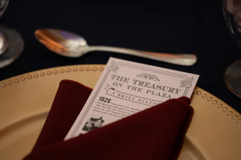 A formal place setting features a cream plate, burgundy napkin, and printed program card with silverware on the table.