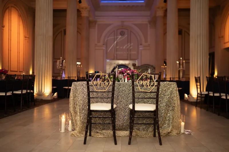 Two black chairs sit at a gold sequined round table beneath tall columns and warm uplighting.