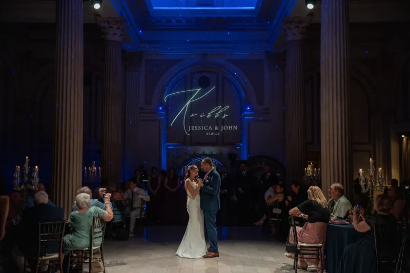 Jessica in white and John in blue share their first dance under blue uplighting.