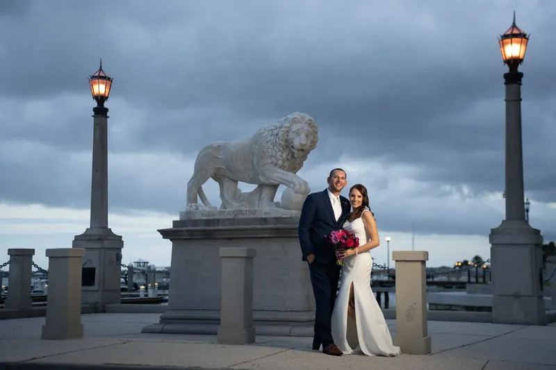 Jessica gazes at John while he looks ahead as they stand together in the columned hallway, her veil trailing behind her.