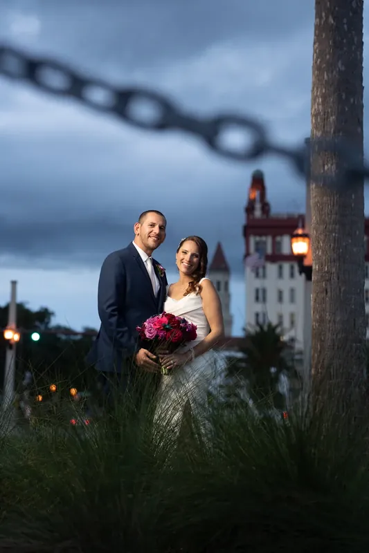 John and Jessica embrace near a weathered post and chain at the bayfront, with historic buildings lit in the background.