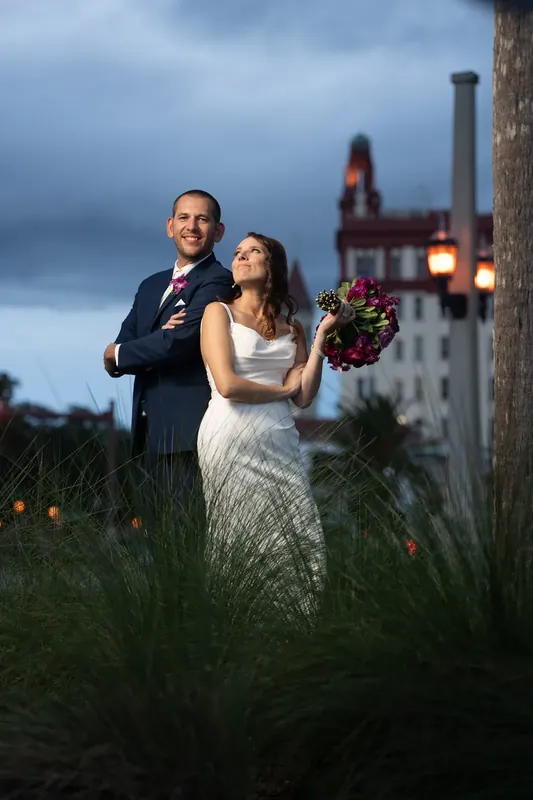 John and Jessica stand close together in tall grass along the bayfront, a historic structure glowing behind them.