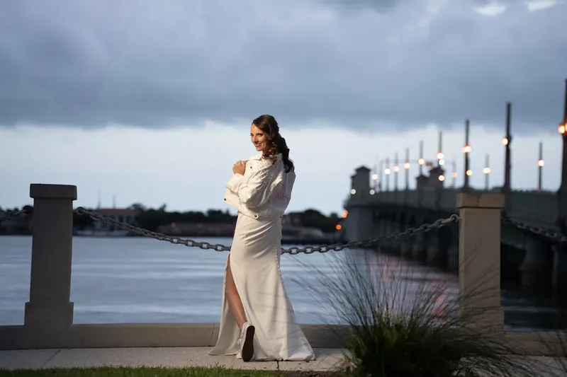 Jessica stands alone in a cream gown, gazing toward the water at the bayfront under gray twilight skies.