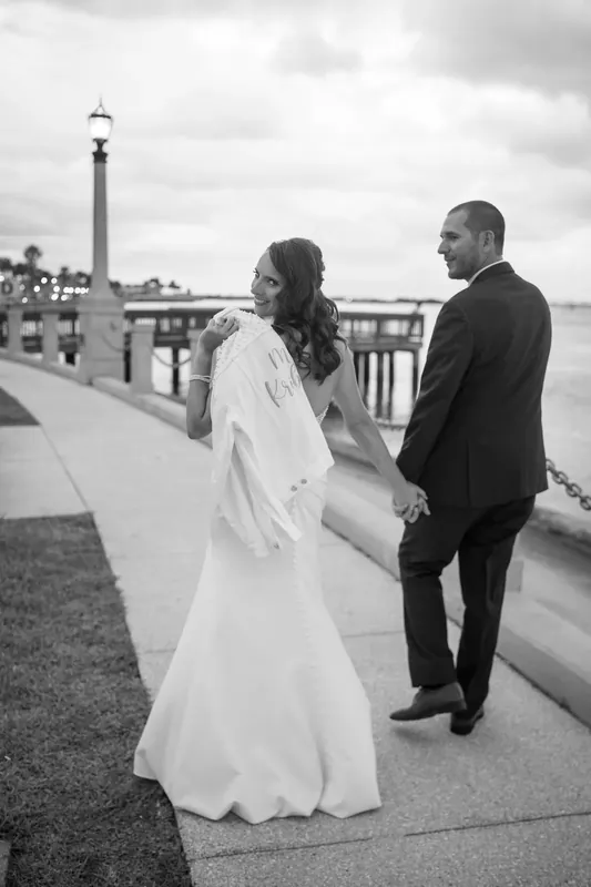 Jessica in a white gown and John hold hands on the bayfront promenade with a lighthouse visible across the water.