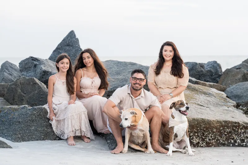 The family crouches on the sand at Vilano Beach, posing with two dogs while massive rocks frame the background.