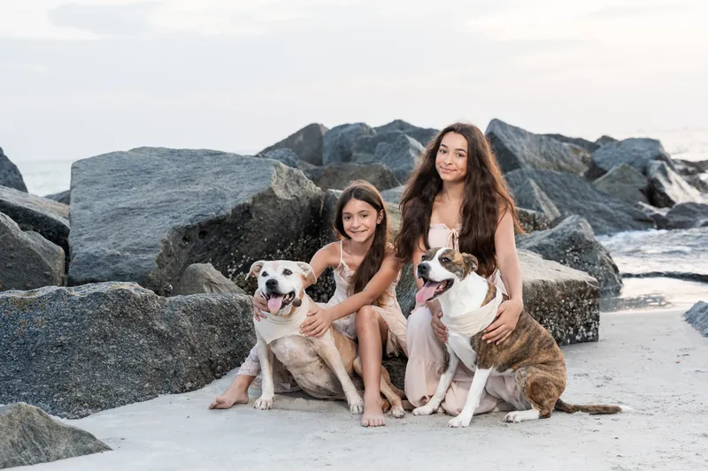 Two of Jessica's daughters sit with two dogs on the beach, smiling at the camera with rocky outcrops behind them.