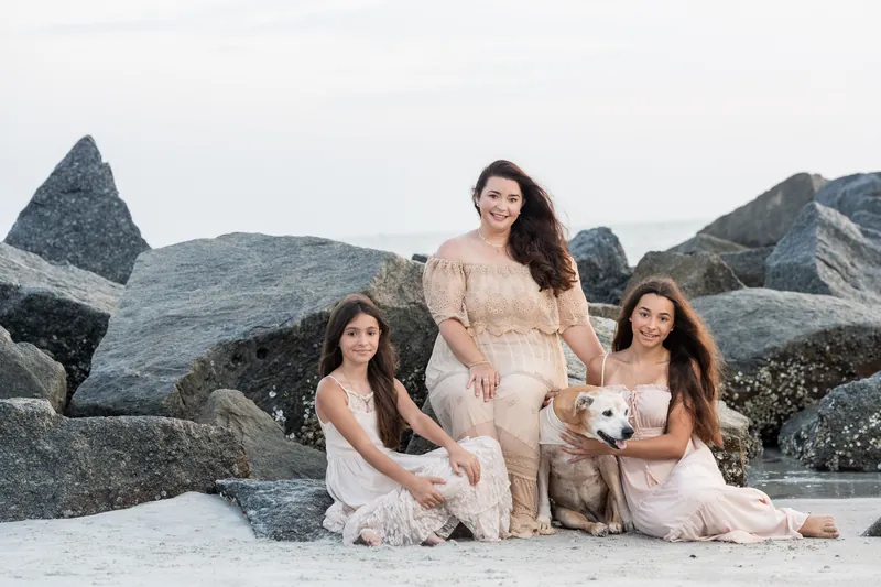 Three females from Jessica's family sit on the sand, wearing cream and beige dresses with boulders rising behind them