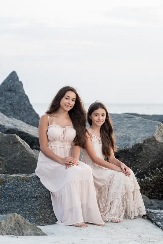 Two members of Jessica's family sit together on the sand, looking toward the ocean with large rocks visible