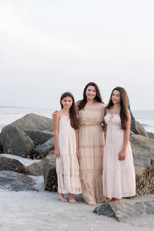 Three females from Jessica's family stand together on the beach in flowing neutral-toned dresses, with boulders and ocean