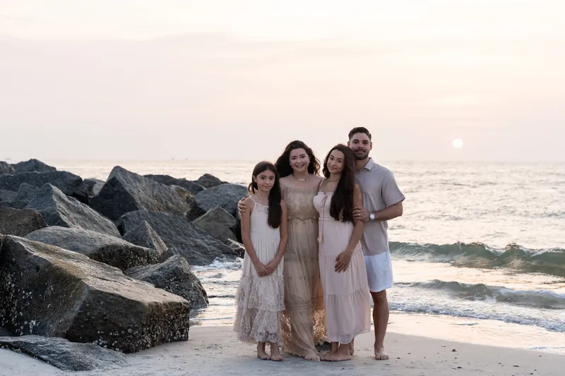 Jessica's family of four stands together on the sand near dark rocks at Vilano Beach, wearing neutral linen clothing