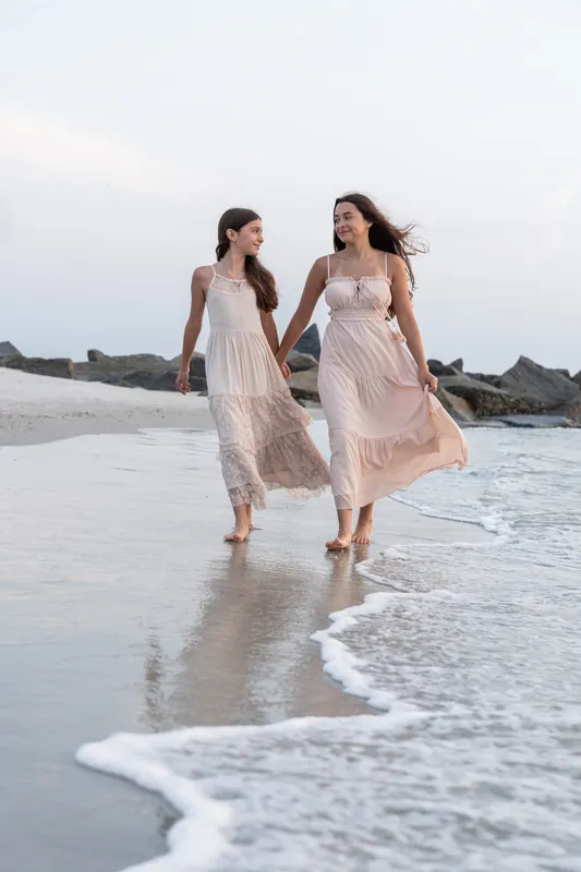 Two young people hold hands and walk through shallow water along the shoreline, their light dresses flowing.