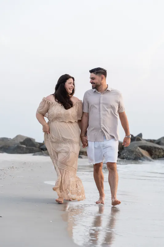 A couple walks along the wet sand holding hands, with large boulders visible behind them along the beach.