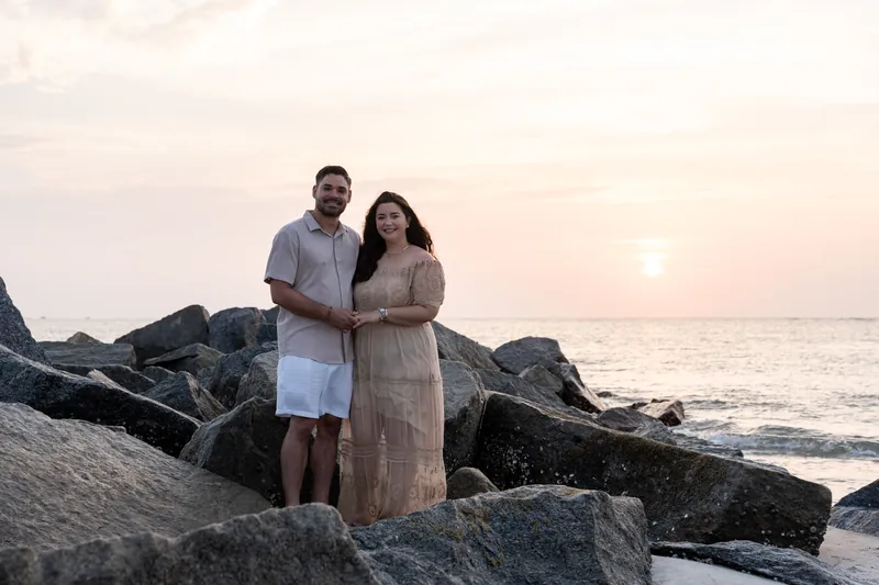 Two people stand close together on dark rocks overlooking the ocean at sunset, wearing soft neutral tones.