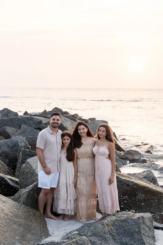 Four family members pose together on the beach rocks with calm ocean waters and pale sky behind them.