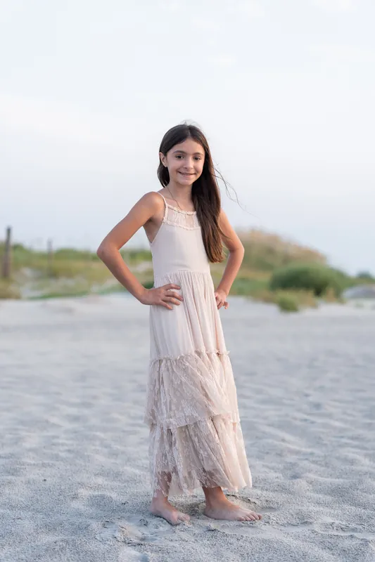 A young person stands alone on the sand with one hand on their hip, wearing a layered cream-colored linen dress.
