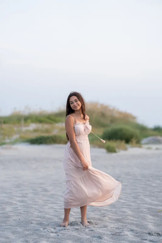 Jessica stands alone on the sand at Vilano Beach in a flowing pale pink dress, smiling at the camera with dunes and beach