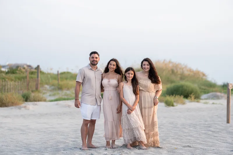 Four family members stand together on the sand wearing neutral tones—beige, cream, and white—with beach dunes visible