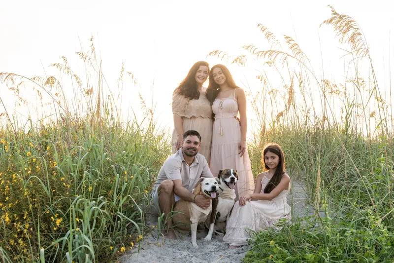 Four family members crouch together among tall beach grasses with a brown and white dog, photographed during golden hour