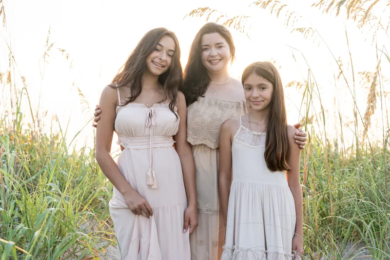 Jessica's family of three stands together among beach grasses at Vilano Beach, wearing soft neutral tones and smiling warmly