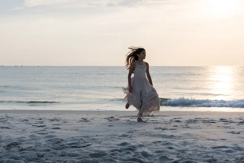 A young woman in a gray dress walks through shallow waves at sunset, her hair flowing as she moves toward the water.