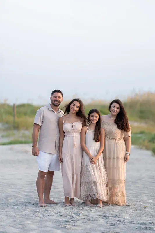 Four family members pose closely on sand, wearing coordinating cream and beige outfits under an overcast sky.