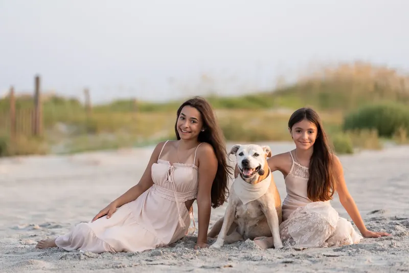Two people sit on sand beside a brown and white dog in a knit wrap, both wearing pale dresses.