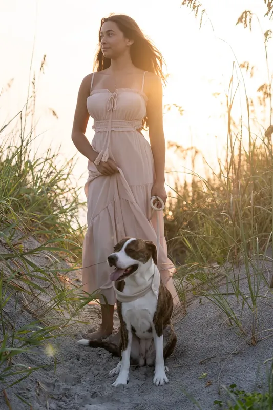 Woman in a light gray sleeveless dress standing with a brindle and white dog at her side among beach grasses at golden hour.