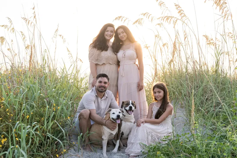 Family of five poses together among tall dried grasses, with two adults and three children in coordinating pale outfits during golden hour.