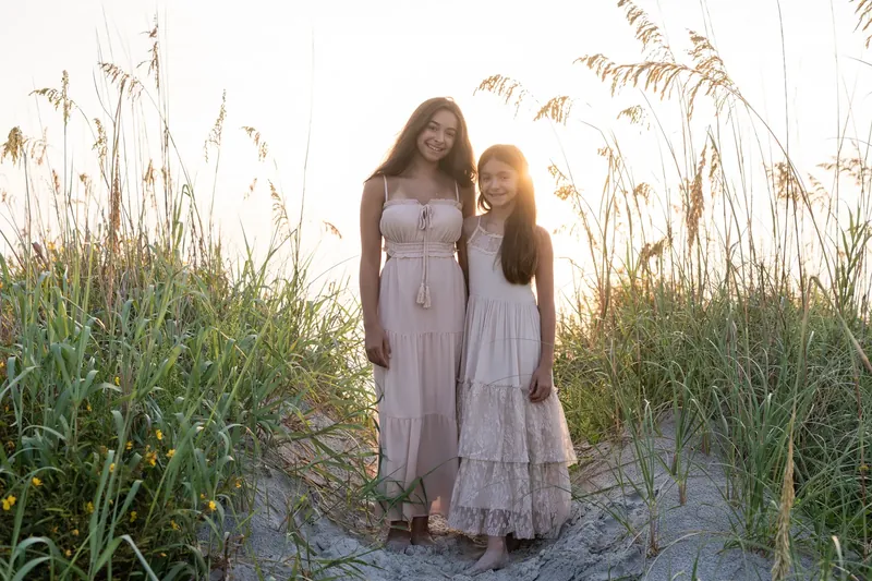 Two people in matching lavender dresses stand close together in a sandy clearing surrounded by beach vegetation at sunset.