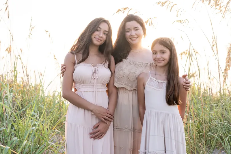 Three females in soft pastels embrace and smile at the camera, framed by tall dried marsh grasses glowing in warm light.