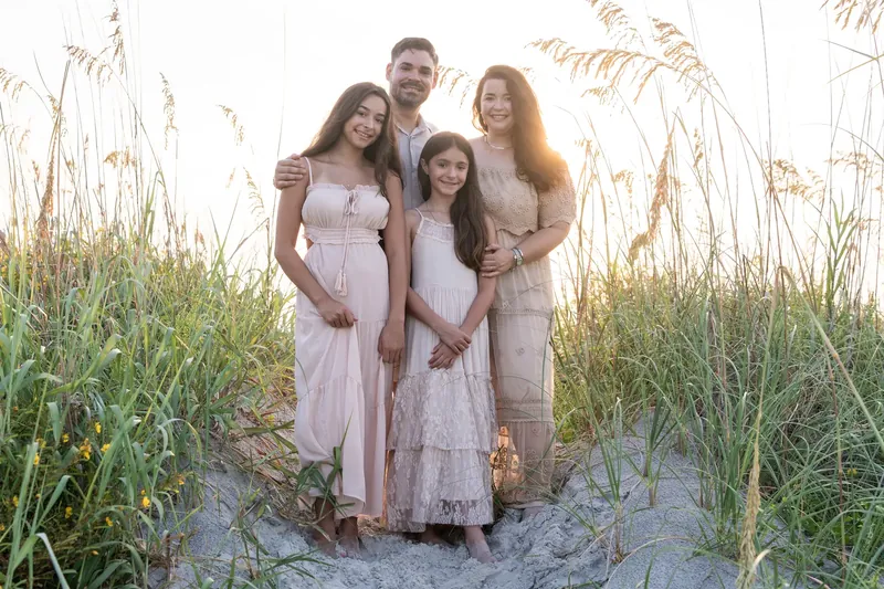 Five family members in coordinated pale clothing gather on sand with dried grasses behind them, smiling at the camera together.