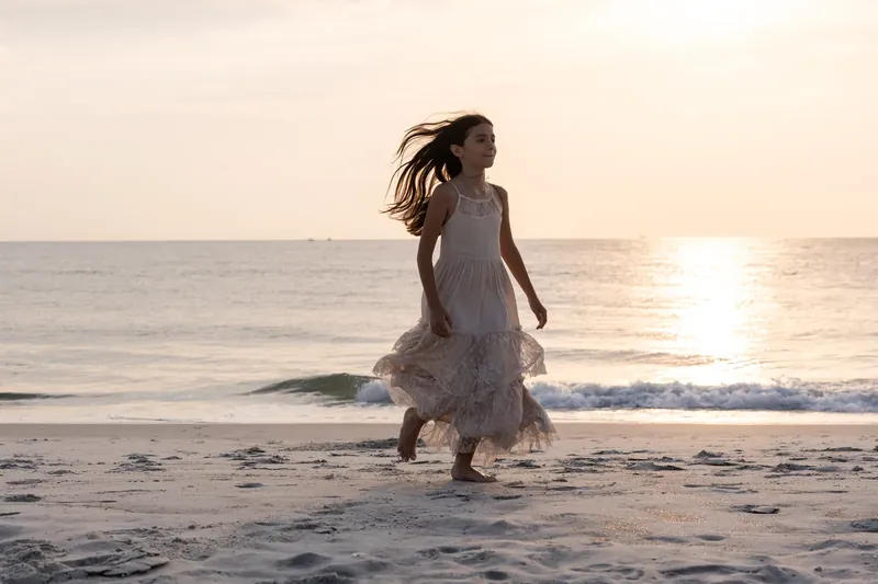 A child in a white dress runs through shallow ocean waves as the sun reflects on the water at dusk.