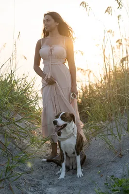 Jessica wears a flowing gray dress and stands on the beach near dune grasses, holding the leash of a brown and white dog
