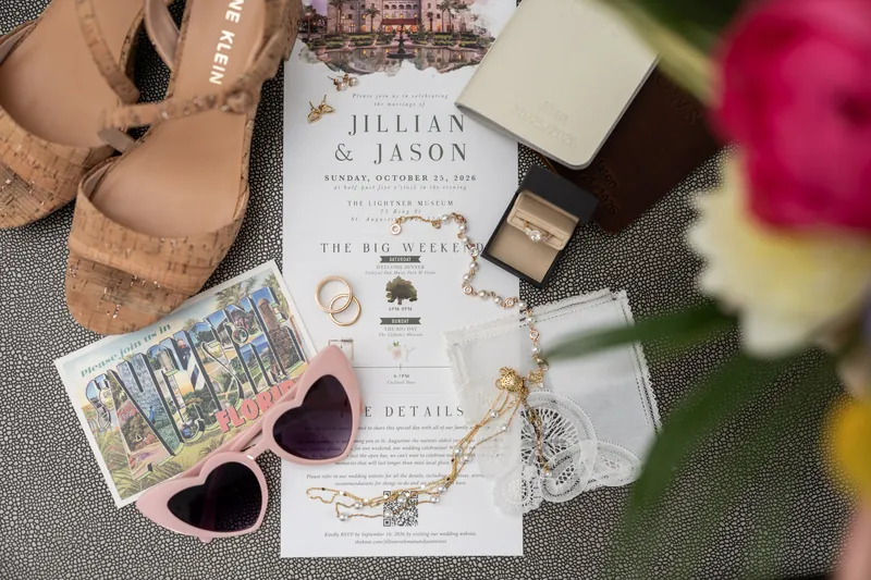 Wedding invitation for Jillian & Jason surrounded by heart-shaped sunglasses, shoes, rings, and flowers on a gray textured surface.