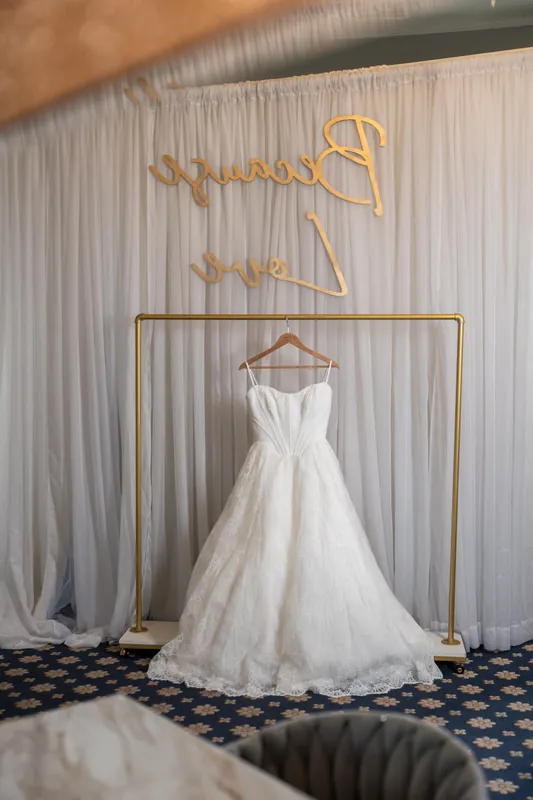 White A-line wedding gown hangs on a gold frame against pleated white curtains with gold wire name signage above at the Lightner Museum.