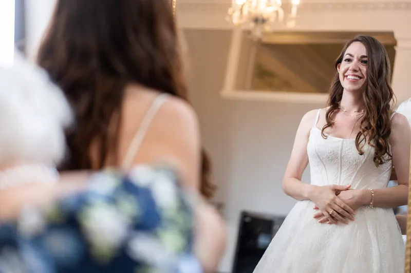 Person in a white sleeveless gown smiles while holding a blue and white bouquet, reflected in an ornate mirror indoors.