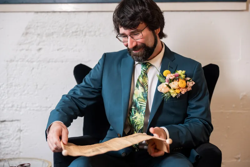 Person in a navy suit with yellow boutonniere examines a rolled document while seated indoors.